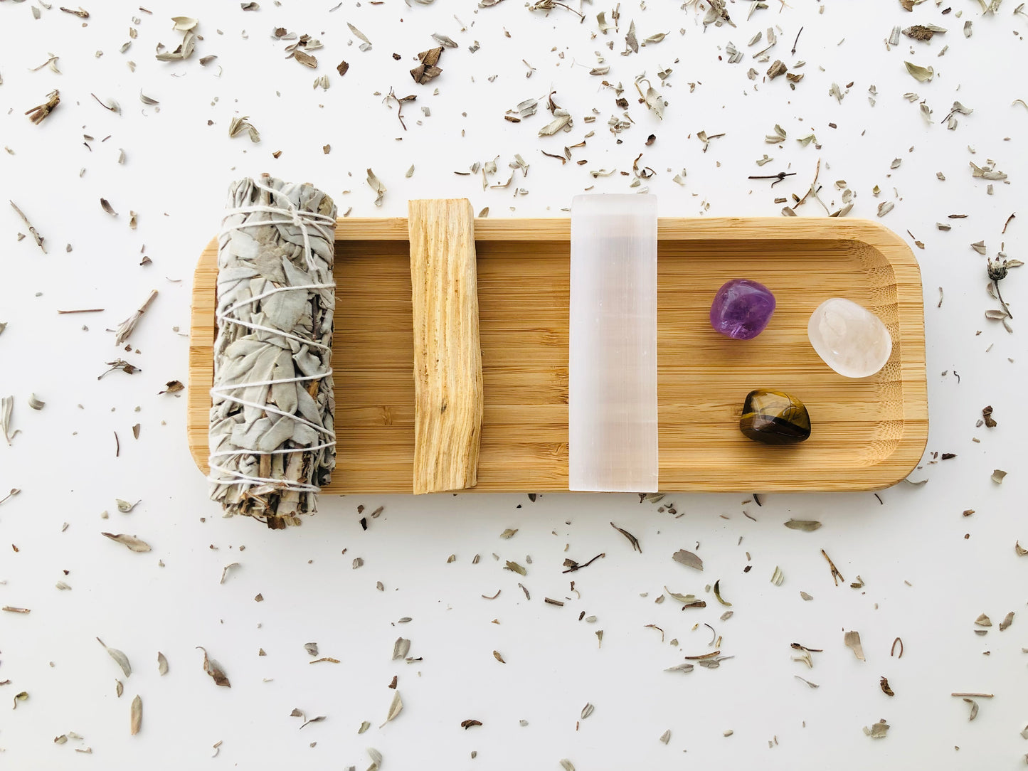 Sobre una superficie blanca llena de polvo de salvia blanca, hay una bandeja rectangular de madera con un atado de salvia blanca, un palo santo, una vara rectangular de selenita y tres rodados de amatista, cuarzo blanco y ojo de tigre.