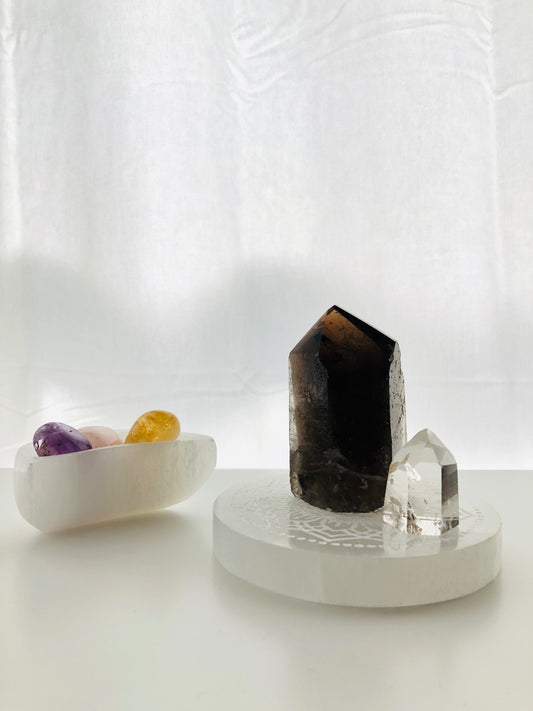 cuenco de Selenita en forma de luna con rodados de citrino, cuarzo rosa y amatista dentro. Al lado, altar redondo de selenita con punta grande de cuarzo ahumado y punta pequeña de cuarzo cristal